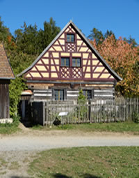 Ein wunderschönes historisches Fachwerkgebäude im Freilandmuseum Oberpfalz. Das Gebäudeensemble wird idyllisch umrahmt von herbstlich gefärbten Bäumen. Ein aus Lärchenholz gefertigter Gartenzaun schützt den bezaubernden Bauerngarten vor ungebetenen Gästen wie Hühner und Gänsen. Die wärmende Herbstsonne lädt den Besucher des Freilandmuseums zum Verweilen ein.